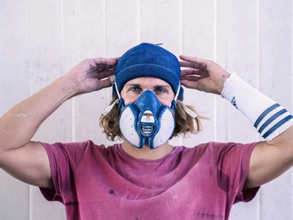Florian Germain, fondateur de FYGOO, face caméra avec masque de ponçage dans son atelier à Capbreton, spécialisé en réparation et finition de planches de surf